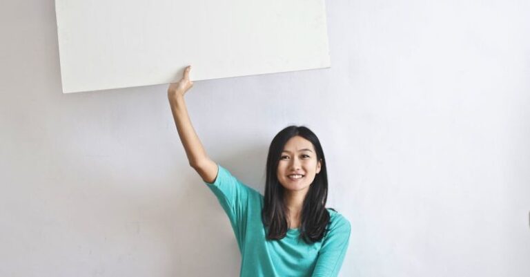 Real Estate Market - Cheerful Asian woman sitting cross legged on floor against white wall in empty apartment and showing white blank banner