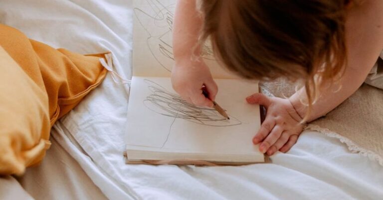 Economic Development - From above of small girl in dress drawing with pencil in notebook while mother using laptop on bed near